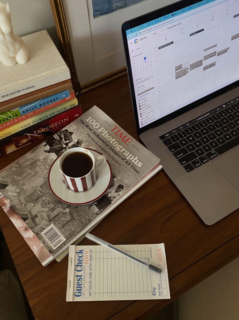 A picture of a macbook computer with a Google calendar open and a coffee sitting on a pile of magazines (next to the laptop).